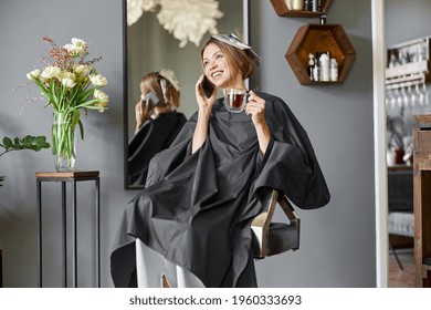 Happy Blonde Caucasian Woman Is Using Smartphone While Sitting In Chair And Waiting In Hairdresser's Salon