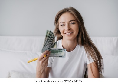 Happy blonde Italian girl in white t-shirt toothy smiles holds dollar banknotes using mine like fan, satisfied by wage. Prize , lottery. Young caucasian female entrepreneur on bed holds cash. Finance. - Shutterstock ID 2254774459