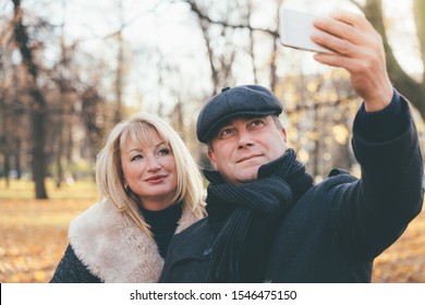 Happy Blonde Mature Woman And Beautiful Middle-aged Brunette Take A Selfie On The Phone. Loving Couple Of 45-50 Years Old Walks In The Autumn Park In Warm Clothes, In A Coat And Enjoys Life