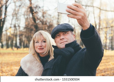 Happy Blonde Mature Woman And Beautiful Middle-aged Brunette Take Selfie On Mobile Phone. Loving Couple Of 45-50 Years Old Walks In The Autumn Park In Warm Clothes, In Coat And Enjoys Life