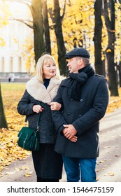 Happy Blonde Mature Woman And Handsome Middle-aged Brunette Man Walk In Park, Looking At Each Other. A Loving Couple Of 45-50 Years Old Walks In The Autumn Park In Warm Clothes