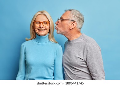 Happy Blonde Middle Aged Woman Gets Kiss In Cheek From Beloved Husband. Senior Couple Have Romantic Relationship In Old Age. Affectionate Male Pensioner Kisses Wife Stand Indoor Over Blue Wall