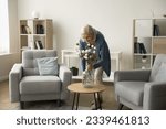 Happy blonde retired mature woman decorating home living room interior, placing flowers in glass bottle vase, smiling, enjoying care of apartment design, floristic hobby