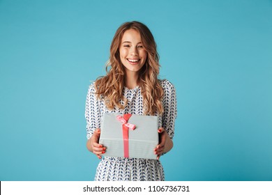 Happy blonde woman in dress holding gift box and looking at the camera while enjoys over blue background