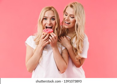 Happy blonde woman eating donut while her sister standing behind her over pink background
