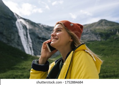 Happy blonde woman hiker in yellow raincoat talks on the phone and smiles on waterfall background in sunny summer day in Norway - Powered by Shutterstock