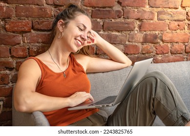 Happy blonde woman with laptop sitting on couch against brick wall. Remote work. High quality photo
