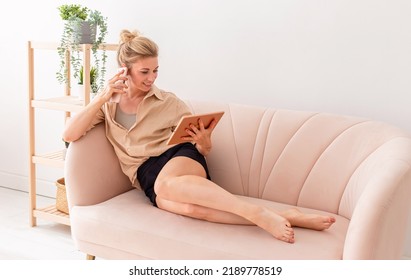 Happy Blonde Woman, Looking At A Digital Tablet And Talking On A Mobile Phone Lying On A Beige Sofa