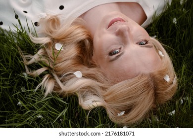 Happy Blonde Woman Lying Down On The Green Grass Enjoying The Summer Season. Close-up Portrait From Above With Natural Makeup