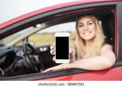 Happy Blonde Woman Showing Smartphone Out The Window Of A Car. Focus On Mobile Phone.