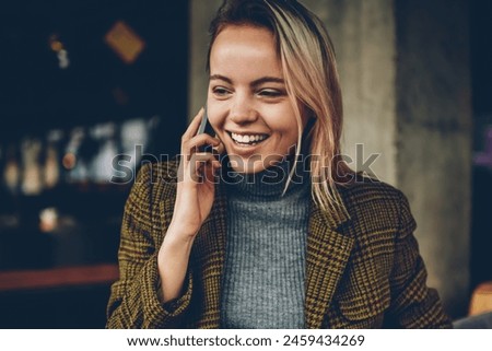 Happy blonde young entrepreneur in elegant jacket calling to best friend and laughing during mobile conversation on smartphone.Positive pretty female blogger talking on telephone resting in cafe