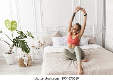 Happy blonde-hair woman is sitting on the bed, stretching after sleep, smiling. Attractive young female in stylish sportswear warm up before workout, morning training concept. High quality photo - Shutterstock ID 1934870519