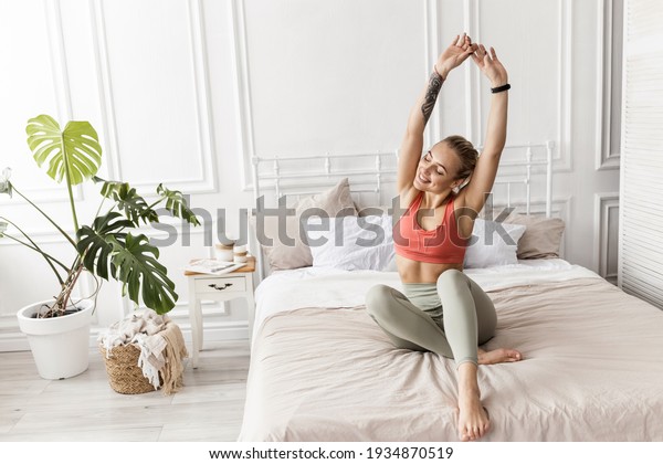 Happy blonde-hair woman is sitting on the bed,\
stretching after sleep, smiling. Attractive young female in stylish\
sportswear warm up before workout, morning training concept. High\
quality photo