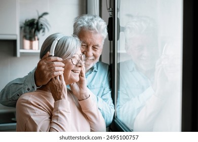 Happy bonding aged senior retired couple standing near window, looking in distance, enjoying peaceful moment together at home. Elderly woman listening to music with headphones - Powered by Shutterstock