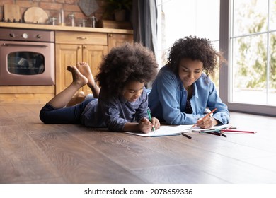 Happy bonding family little African ethnicity child girl involved in creative activity with happy young biracial mother, drawing pictures in paper album lying together on heated floor.