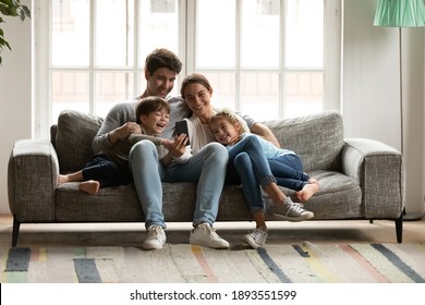 Happy bonding family with two adorable small kids looking at cellphone screen, laughing at funny video or photos in social networks, holding distant video call with grandparents, having fun at home. - Shutterstock ID 1893551599