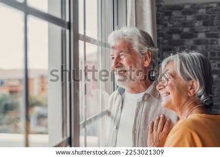Happy bonding loving middle aged senior retired couple standing near window, looking in distance, recollecting good memories or planning common future, enjoying peaceful moment together at home.