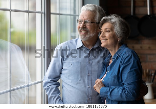 Happy bonding loving middle aged senior retired\
couple standing near window, looking in distance, recollecting good\
memories or planning common future, enjoying peaceful moment\
together at home.