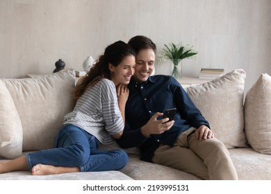 Happy Bonding Millennial Hispanic Family Couple Using Mobile Applications, Relaxing On Cozy Sofa, Playing Online Games On Cellphone, Watching Funny Photo Video Content, Modern Tech Addiction Concept