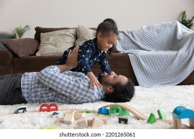 Happy Bonding Young African American Father Playing With Cute Little Kid Daughter, Lying On Floor Carpet In Modern Living Room. Joyful Laughing Two Generations Family Having Fun On Weekend At Home.