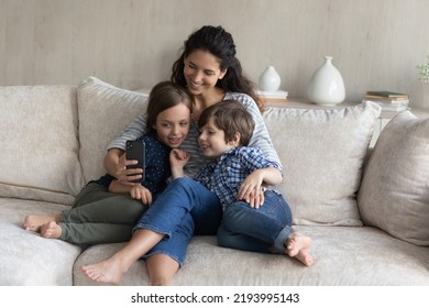 Happy Bonding Young Latin Mum And Cute Kids Siblings Posing For Selfie Photo On Cellphone Or Recording Streaming Live Video Stories In Social Network, Resting Together On Sofa, Tech Addiction.