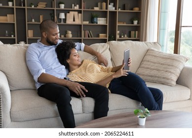 Happy Bonding Young Married Mixed Race Family Couple Using Digital Tablet, Planning Travel Booking Flight Tickets, Watching Video Content In Social Network, Shopping Online, Resting On Couch At Home.