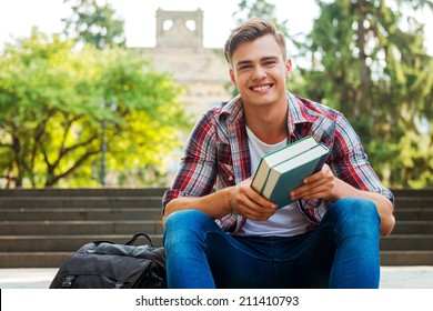 Happy bookworm. Handsome male student holding textbooks and smiling while sitting at the outdoors staircase with university building in the background - Powered by Shutterstock