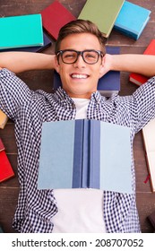 Happy Bookworm. Top View Of Happy Young Man Holding Hands Behind Head And Smiling While Lying On The Hardwood Floor With Colorful Books Laying All Around Him 