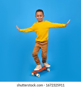 Happy boy is African American riding a red skateboard balancing his arms placed in sides for a balance-clad yellow sweater and brown pants against a blue studio background. free space for text - Shutterstock ID 1672325911