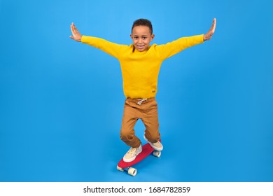 Happy boy is African American riding a red skateboard balancing his arms placed in sides for a balance-clad yellow sweater and brown pants against a blue studio background. free space for text - Shutterstock ID 1684782859