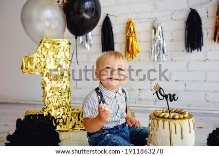 Happy boy blonde 1 year old on his birthday tries sweets. The photo zone with the first cake is decorated in black and gold. Crashcake