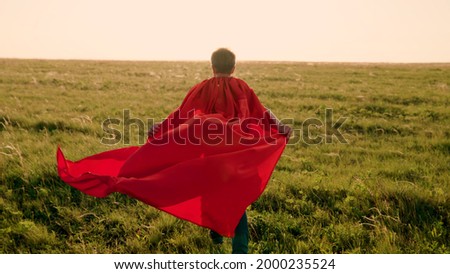 Happy boy, child superhero, runs across green field in red cloak, cloak flutters in wind. Childrens games, dreams. Slow motion. Teenager dreams of becoming superhero. Red cloak jerk, dream expression