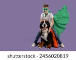 Happy boy with cute fluffy dog in superhero costumes on purple background