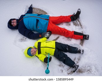 Happy Boy And Dad In Colorful Winter Clothes Doing Gymnastics On Ice And Snow. Father With Child Is Playing Outdoors In Snow. Outdoor Fun For Winter Holidays. Snow Angels