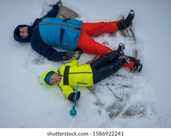 Happy Boy And Dad In Colorful Winter Clothes Doing Gymnastics On Ice And Snow. Father With Child Is Playing Outdoors In Snow. Outdoor Fun For Winter Holidays. Snow Angels