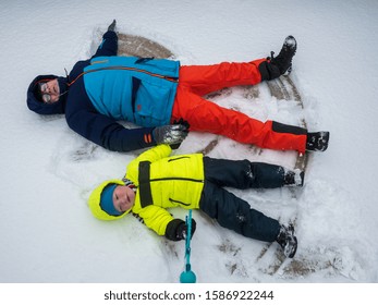 Happy Boy And Dad In Colorful Winter Clothes Doing Gymnastics On Ice And Snow. Father With Child Is Playing Outdoors In Snow. Outdoor Fun For Winter Holidays. Snow Angels