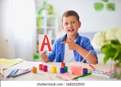 Happy boy does his homework at home. Cute child at table with school supplies smiles funny and learns the alphabet in a playful way. positive student in a bright room with painted letters in his hands - Shutterstock ID 1683222010