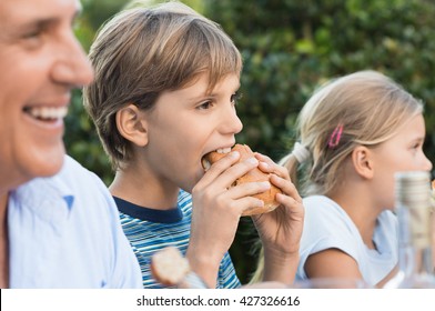 Happy Boy Eating Hamburger With His Family. Young Boy Having Lunch With Sandwich. Portrait Of A Child Eating At Family Party.