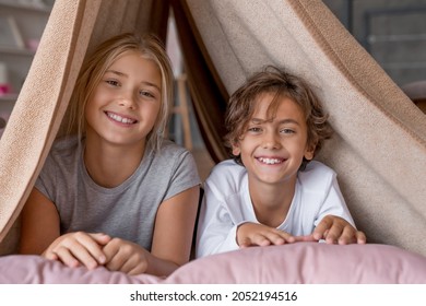 Happy boy and girl sitting in fort from blankets and smiling in camera. Siblings sister and brother playing together on the bed in the morning.