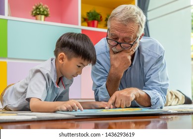 Happy Boy Grandson Reading Book With Old Senior Man Grandfather At Home