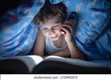 Happy Boy Is Lying Under Blanket And Reading Book In Dark With Flashlight, Instead Of Sleeping, At Time When Everyone Is Asleep, Night. Kid Hid From Everyone. Thirst For Knowledge.