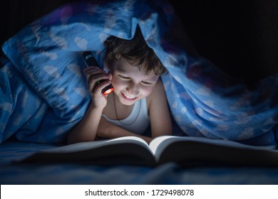 Happy Boy Is Lying Under Blanket And Reading Book In Dark With Flashlight, Instead Of Sleeping, At Time When Everyone Is Asleep, Night. Kid Hid From Everyone. Thirst For Knowledge.