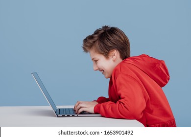 happy boy n red sweatshirt playing on laptop, iisolated blue background, profile view