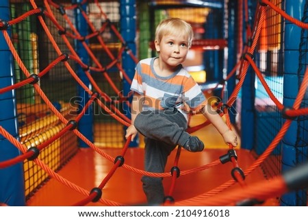 Happy boy playing in indoor playground on net trail. Child having fun on modern playground. Cute kid playing on colorful playground at shopping mall