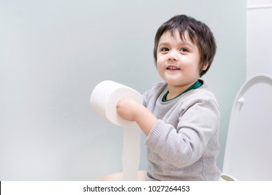 Happy Boy Playing With  Toilet Paper In Toilet,Healthy Kid Sitting With Holding A Roll Of Toilet Paper And Lookign At Camera With Smiling Face,Child Tear Toilet Roll Against Light Green Background