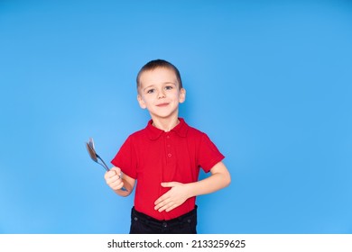 The Happy Boy Points To His Full Stomach And Holds A Fork And Spoon In His Hand On A Blue Background. The Concept Of Proper Nutrition.