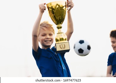 Happy Boy Rising Golden Trophy. Child Winning Sports Competition. Overjoyed Kid Holding Golden Prize Award. Soccer Ball In The Background