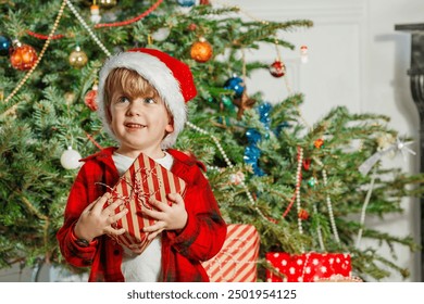 A happy boy with a Santa hat holds onto some presents on Christmas Eve a beautifully decorated xmas tree in the background - Powered by Shutterstock