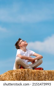 Happy Boy Sits On Haystack Against Blue Sky. Child Enjoys The Fresh Air Looking Up To The Sky In The Village. Vertical Frame.