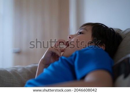 Happy boy sitting on sofa bitting finger nails while watching cartoon on TV. Mixed race Child resting in living room with bright light shining from window on sunny day summer. Kid relaxing at home